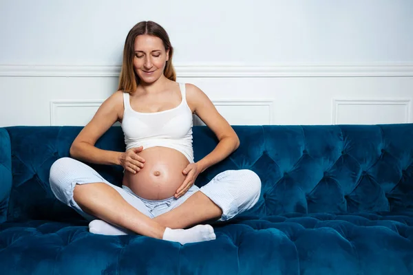 幸せな肖像画の妊娠中の女性座っています上のソファ冷静に腹に触れて — ストック写真