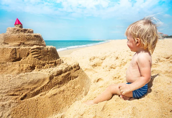 Szczęśliwy Chłopczyk Siedzi Przed Piaskowym Zamkiem Patrzy Morze Plaży — Zdjęcie stockowe