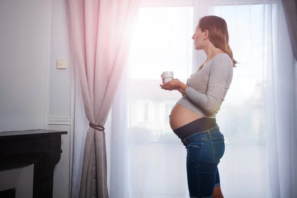 一个孕妇站在窗边喝茶看外面的轮廓 — 图库照片