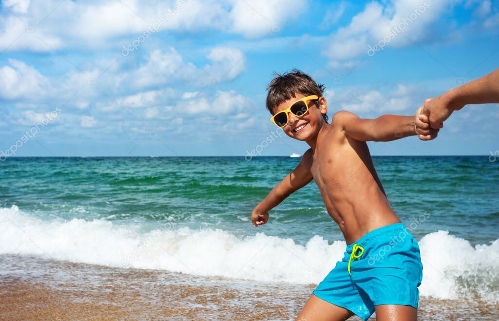 Happy boy in sunglasses pull parent's hand to the ocean smiling going to the sea water waves