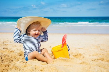 Küçük mutlu çocuk plajda oturur büyük hasır şapka takar ve plastik kovayla oynar.