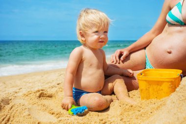 Mutlu küçük bir çocuğun portresi deniz kenarındaki kumda hamile annenin karnına yakın oturuyor.