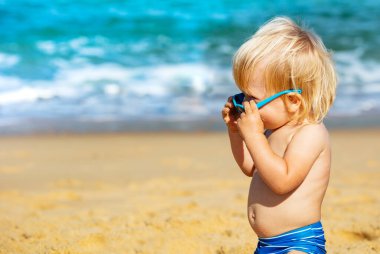 Mutlu küçük çocuk sahilde sürünüyor, koruyucu güneş gözlüğü takıyor, harika bir bakış atıyor.