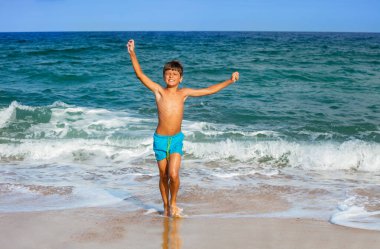 Güneş gözlüklü, mutlu ve rahat bir çocuğun portresi. Kocaman gülümseyen, deniz dalgalarında koşan elleri kaldıran.