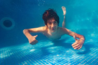 Havuzda gülümseyen bir portreyle suyun altında yüzen mutlu yakışıklı çocuk baş parmağını kaldırıp el hareketi yapıyor.