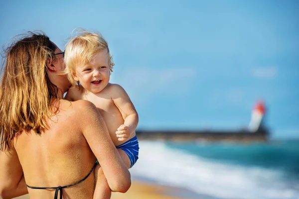 笑顔の幼児の男の子の幸せな肖像画を笑って見て 背景に灯台と海の近くのビーチに立っている母親の肩の上に笑う — ストック写真