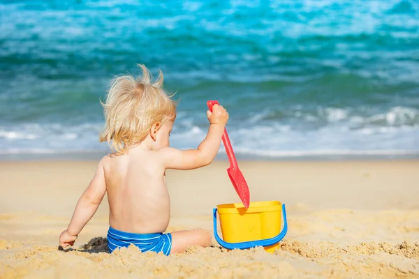 小さな幸せなブロンド男の子遊びますともに小さなバケツホールドシャベルでザビーチ近くの海の景色からバック — ストック写真