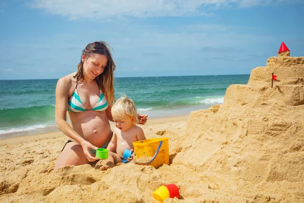 Szczęśliwa Ciężarna Matka Siedzieć Plaży Małym Maluchem Blond Chłopiec Pobliżu — Zdjęcie stockowe