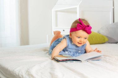 Little baby girl with beautiful red bow read book bed in nursery wearing dress