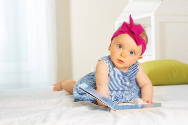 Infant baby girl with curious look and beautiful red bow laughing while looking, leaf through book bed in nursery wearing dress