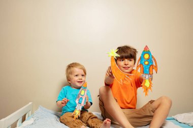 Two happy brothers play with rockets drawn on paper on the bed together smiling clipart