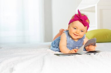 Cute tiny baby girl with curious look and beautiful red bow laughing while leafing through book bed in nursery wearing dress
