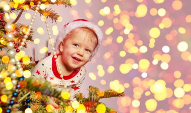 Happy smiling little toddler boy in Santa hat looking from behind of the Christmas tree