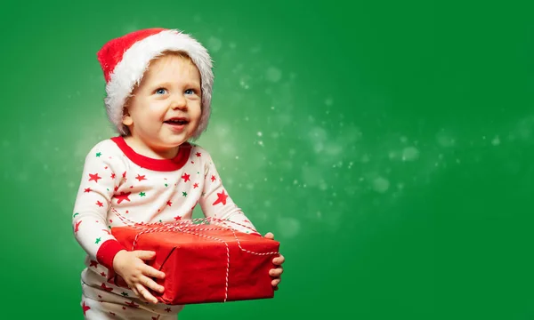 可爱的蹒跚学步的男孩拿着礼物盒和绿色背景雪地的圣诞老人帽 — 图库照片