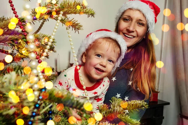 幸せな小さな幼児男の子とともにお母さん見て笑顔後ろクリスマスツリー身に着けているサンタ帽子 — ストック写真