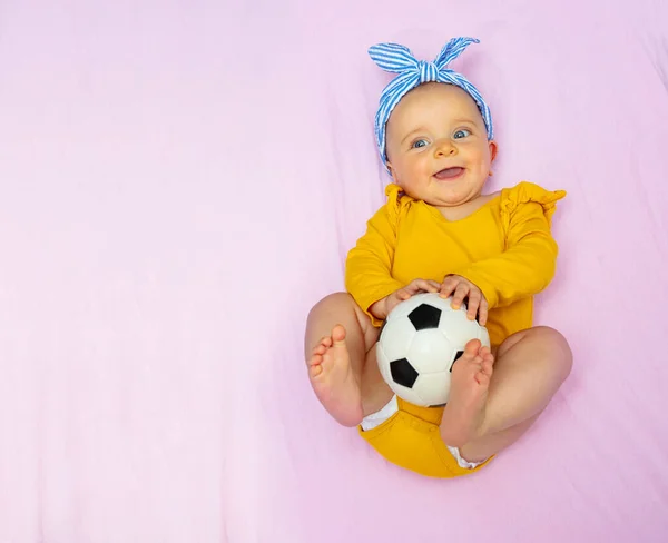 笑顔の小さな赤ちゃんの女の子を保持サッカーボールはベッドの着用弓に横たわり 感動的な足を見上げます 上からの眺め — ストック写真