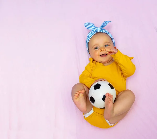 笑い幸せな小さな赤ちゃんの女の子ホールドサッカーボールレイでベッドウェア弓とルックアップ感動的な脚 上からの眺め — ストック写真