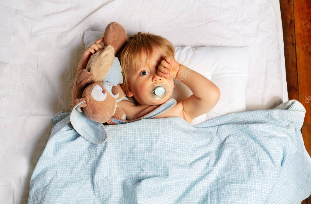 Sleeping baby boy waking up in the bed hugging toy and wipe eyes