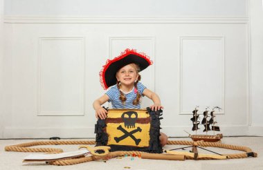 Korsan şapkası takan bir kız çocuğu, yanında model gemi gibi nesnelerle bir sandığın yanında görülüyor.