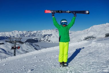 Neon yeşili pantolon giyen bir kayakçı, rüzgarlı bir yamaçta kayakları kaldırır. Mavi gökyüzünün altına uzanan pürüzlü karlı zirveler.