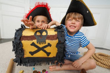 Korsan kostümlü iki oyuncu çocuk sevinçle sunar. Kafatası ve çapraz kemikle işaretlenmiş bir korsan kutusu. Şekerler ve küçük gemilerle çevrili.