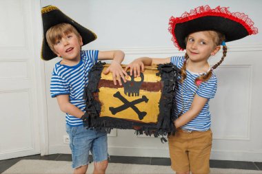 İki kardeş, çizgili korsan gömlekleriyle birlikte sırıtırken ağır hazine sandığının yükünü paylaşıyor.