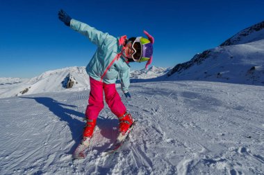 Güneşli bir dağda, renkli giysiler içinde genç bir kayakçı sevinçle bir tarafa eğilir karla kaplı zirvelere karşı.