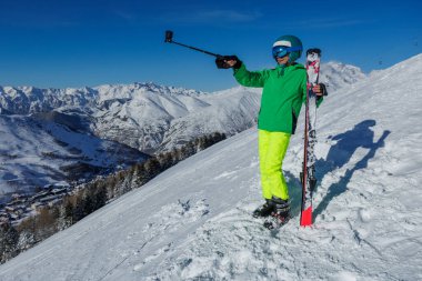 Parlak yeşil ceketli bir kayakçı, arkasında dağ manzaraları olan karla kaplı bir tepenin üstünde selfie çubuğu ve kayak tutuyor.