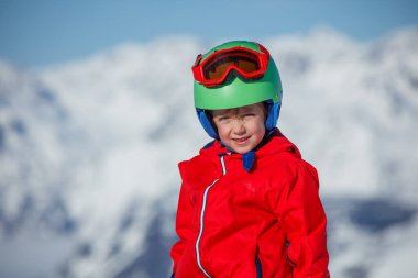 Kırmızı ceketli ve yeşil kasklı gülümseyen bir çocuğun yakın çekimi, güneşli bir günde karlı dağlar yavaşça bulanıklaşır.