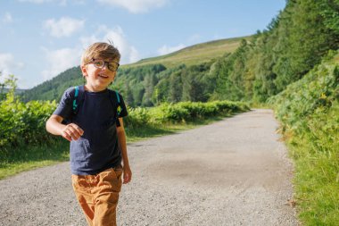 Gülümseyen, gözlüklü bir çocuk, bitkilerle dolu kırsal bir yolda yürürken güneşli bir günün tadını çıkarır.