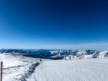Bulutsuz mavi gökyüzünün altında uzanan karlı dağlar. Kıtır, beyaz kar altında görünen kayak yolları.