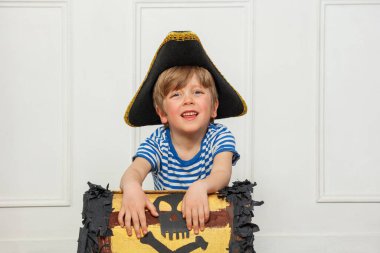 Korsan gibi giyinmiş bir çocuk, yanında deniz temalı eşyalar olan bir hazine kutusunun yanında oturuyor.