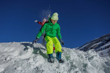 Üç çocuk karda heyecanlı bir şekilde zıplıyorlar, arkalarında açık mavi gökyüzüyle yamaçtan aşağıya