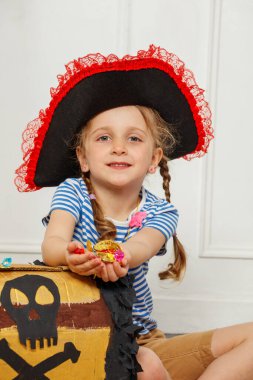 Üzerinde kafatası amblemi olan bir hazine kutusunun yanında oturan korsan şapkalı bir kız bir avuç parlak mücevheri kavrıyor.