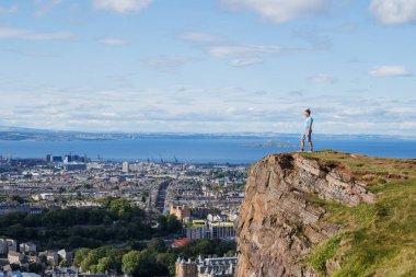 Kayalık bir uçurumun kenarında duran bir insan, şehir manzarasına ve okyanusa, parlak bir gökyüzünün altında bakar.