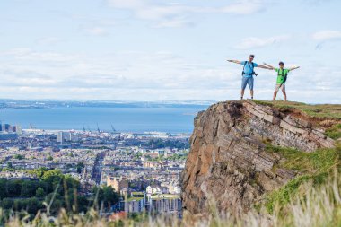 Kalabalık bir kasabanın ve geniş suların üzerinde iki gezgin, Arthurs Koltuğu, Edinburg 'daki bir kayalığın tepesinde kollarını uzatarak tırmanışlarını kutluyor.