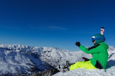 Yeşil ceketli bir genç kayakçı, elinde bir kamerayla güneşli dağ ufkuna bakarken dik bir şekilde fotoğraf çekiyor.