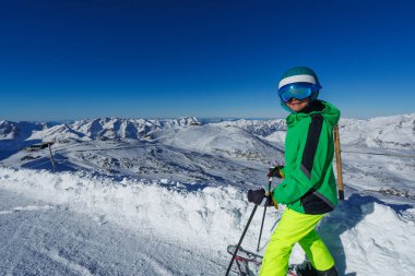 Güneşli bulutsuz bir günde karlı dağlarla renkli giysiler içinde kayak yapan mutlu genç çocuk.