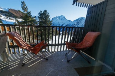 Karlı dağlar ve ağaçlardan oluşan bir zemin ile, iki turuncu sandalye dağ kulübesi verandasına kurulur.