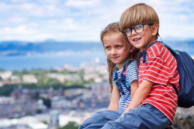 İskoçya Ulusal Anıtı 'nın bir kaya parçasında dinlenen bir kız ve erkek çocuğu, mavi gökyüzü olan bir şehrin panoramik manzarasına hayran.