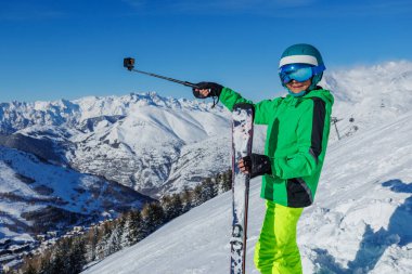 Neon yeşili ceket giyen bir kayakçı el değmemiş bir yamaçta duruyor, taze baruttan güneş parıldarken uzak sivri tepeleri işaret ediyor.