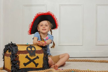 Korsan gibi stili olan ve pırıl pırıl mücevherleri olan bir kız, kafatası resmi olan bir hazine kutusunun yanında oturuyor.