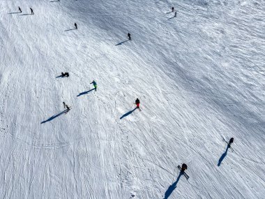 Yüksek açılı bir çekim, kayakçıları taze beyaz kar üzerindeki renkli noktalar olarak gösteriyor. Uzun gölgeleri onları parlak kış yamacında takip ediyor.