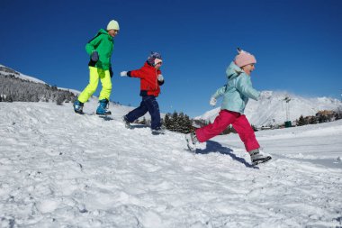 Genç bir çocuk ve iki küçük çocuk karlı dağlarda yokuş aşağı koşuyorlar. Kış tatillerinde dışarıda oynamaktan memnunlar.
