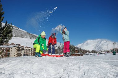 Eğilerek, iki oğlan, bir kız eldivenle taze kar toplar ve onu çam ağaçlarının, uzak tepelerin altına fırlatır.