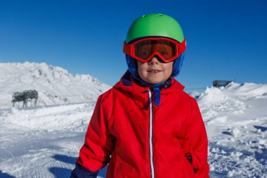 Parlak tebessümlü bir çocuk kırmızı ceketli ve yeşil kasklı, yoğun bir kobalt gökyüzünün altında karla kaplı dağlara bakıyor.