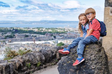 İki çocuk Edinburg 'daki Calton Hill parkında bir kayaya tünemiş parlak bir gökyüzünün altında manzaranın tadını çıkarıyorlar.