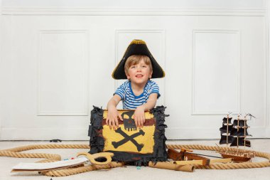 Genç bir çocuk korsan şapkası takıyor ve etrafı deniz aksesuarlarıyla çevrili bir hazine sandığıyla oturuyor.