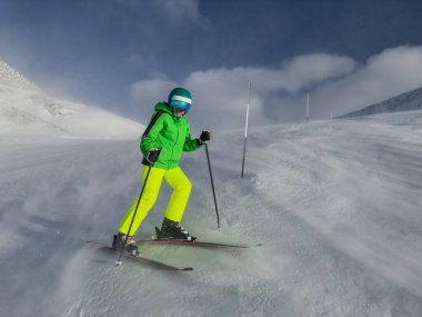 Yeşil kıyafetli genç çocuk karlı, rüzgarlı bir dağ yamacında kayak yapmayı sever. Arka planda bulutlar.