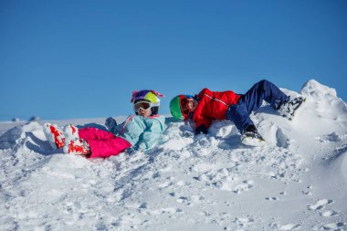 Renkli ceketler içinde iki çocuk, kar yığınına uzanmış, açık gökyüzünün altındaki el değmemiş beyaz yamaçta dinlenirken sırıtıyorlar.